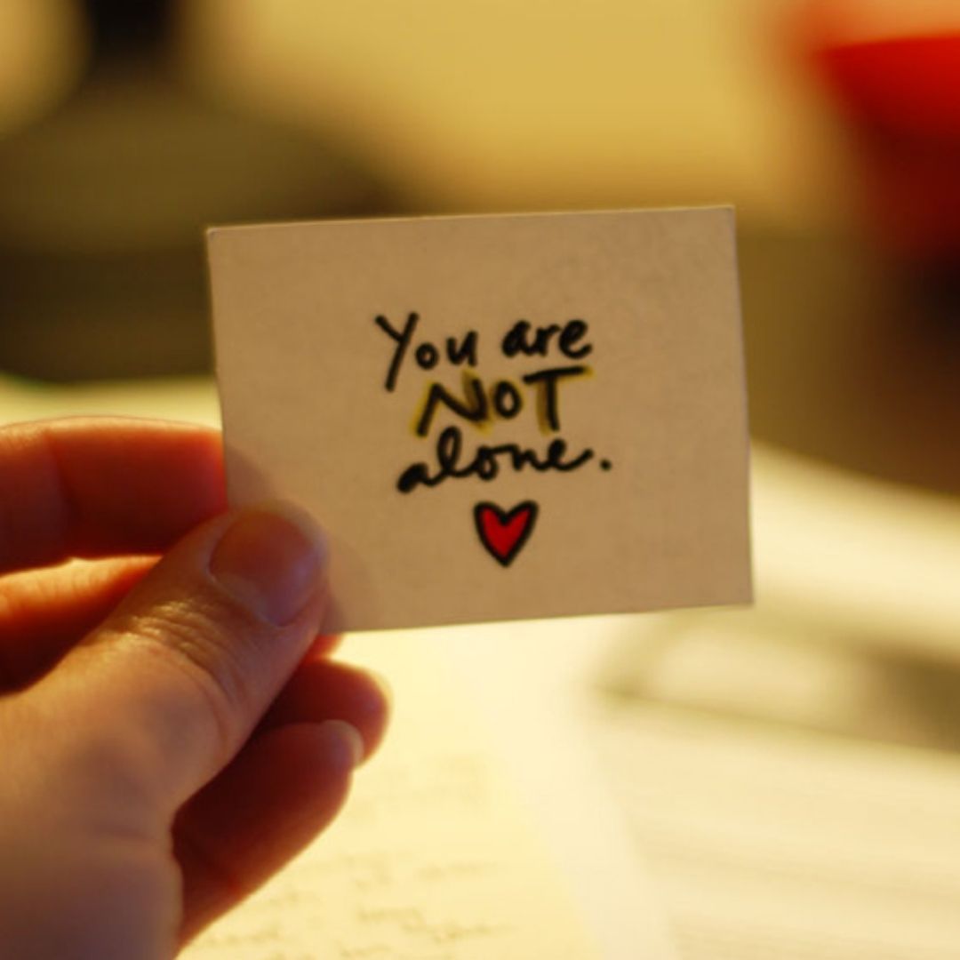 A person's hand holding out a small square sheet of paper which says "You are not alone".